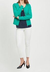 Zielona kurtka o fakturowanym wykończeniu, granatowa bluzka pod spodem, białe slim-fit spodnie i czarne wysokie obcasy. Zaprojektowana z okrągłym dekoltem i zapięciem na zamek.