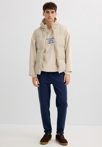 Chaleco acolchado beige con cuello alto sobre una sudadera con capucha beige claro con texto azul, combinado con pantalones deportivos azul marino y zapatillas marrones.