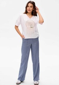 Weißes kurzärmliges Hemd mit V-Ausschnitt und goldenem Baumdesign, kombiniert mit hellblauen weit geschnittenen Hosen und weißen Turnschuhen.