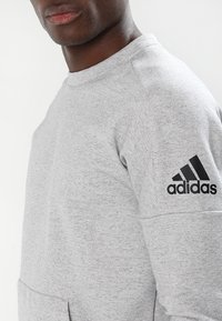 Sweatshirt gris en tricot avec un col rond, un tissu texturé et un logo Adidas noir sur l'épaule gauche.