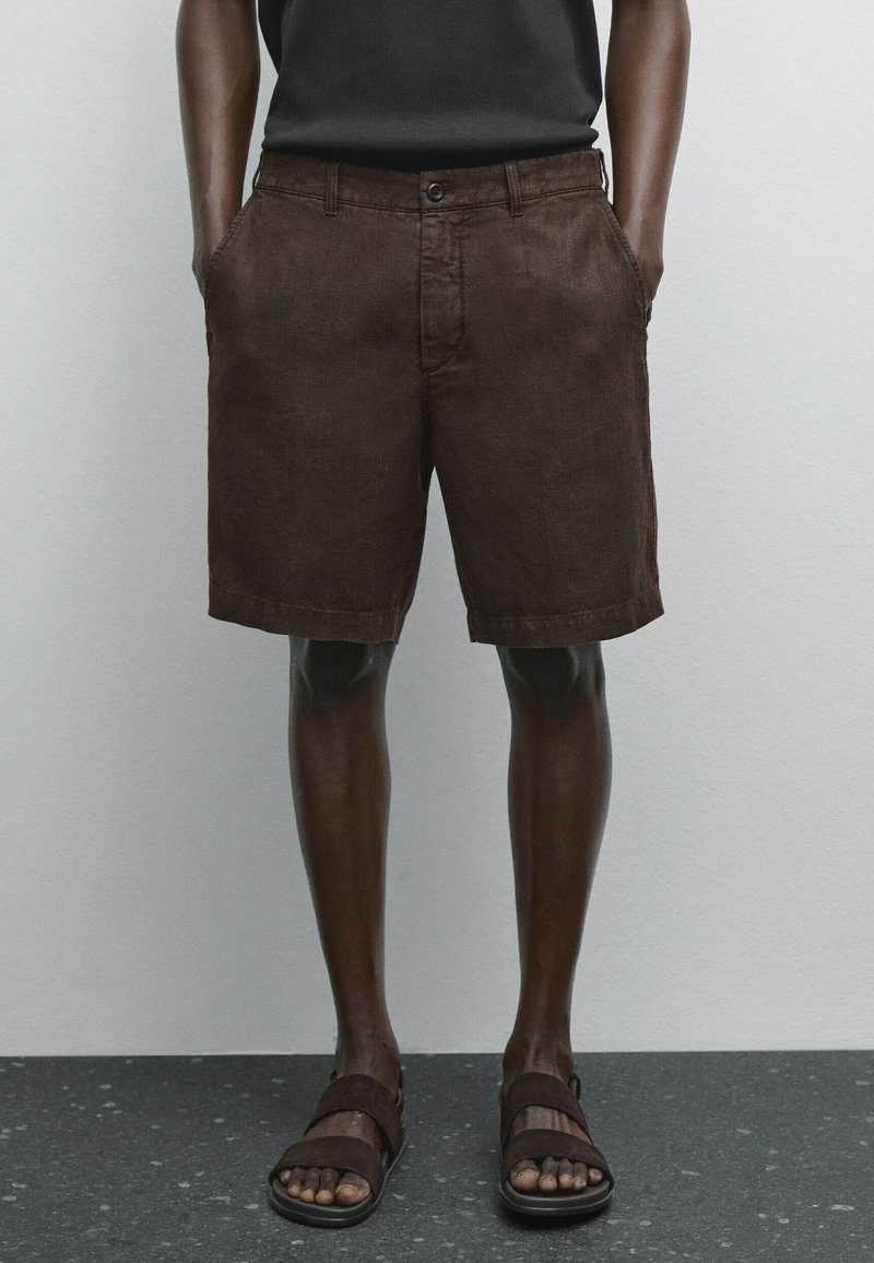 Bruna linnebyxor med sidofickor, knäppning och fållade kanter, kombinerat med mörkbruna sandaler på nakna fötter.