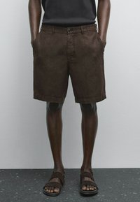 Shorts en lin marron avec poches latérales, fermeture à bouton, bords ourlés et coupe décontractée, associés à des sandales marron sur une surface sombre.