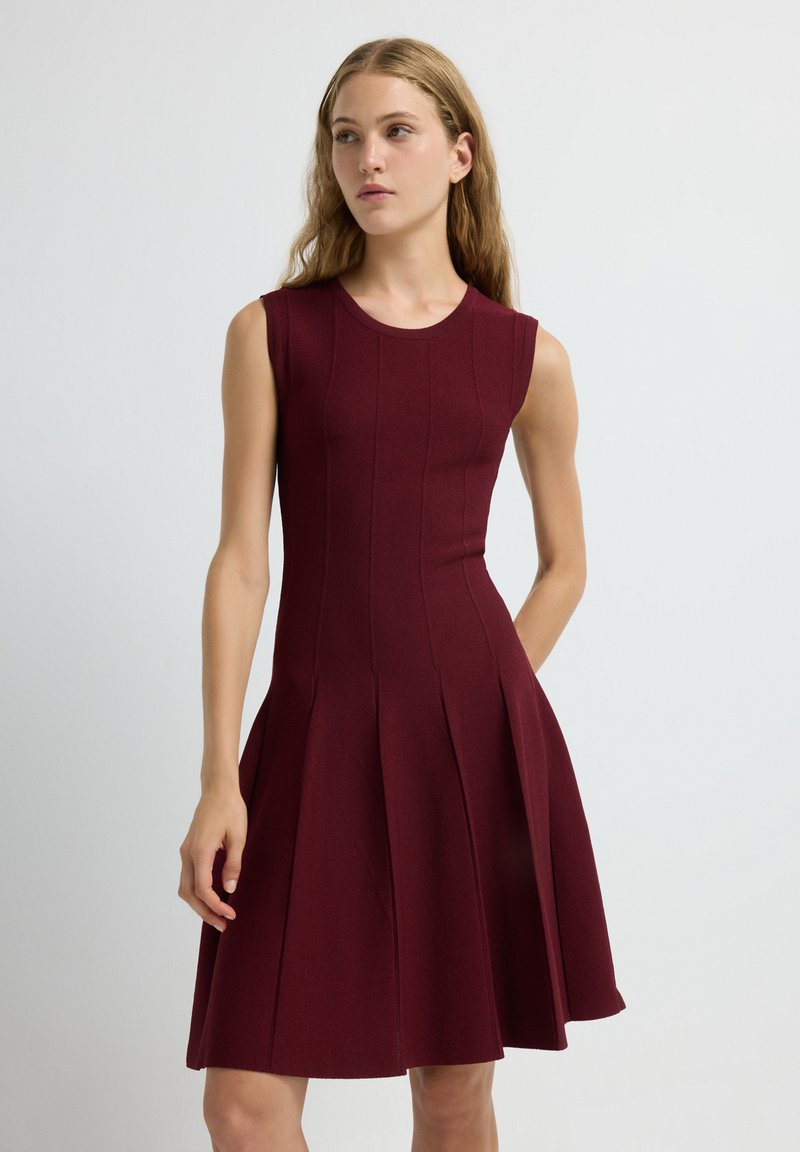 Robe sans manches bourgogne avec un corsage ajusté et une jupe évasée, plissée. Texture lisse, détails à nervures et col rond.