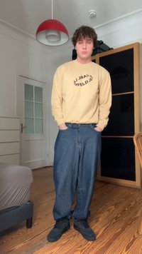 Camicia beige a maniche lunghe con testo circolare nero, abbinata a jeans blu larghi. Sneakers nere. Sfondo con pavimento e mobili in legno.