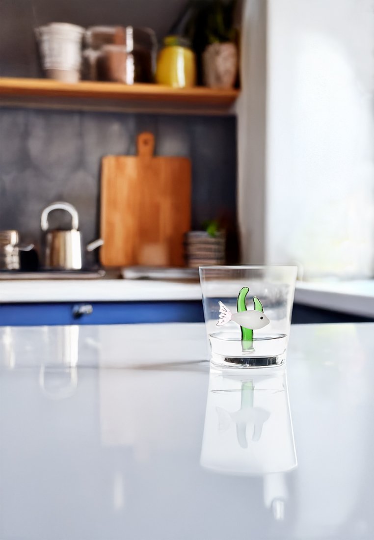 Szkło z wodą i dekoracyjnym wzorem ryb oraz wodorostów na białym blacie kuchennym, rozmyte tło z czajnikiem i deską do krojenia.