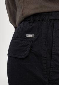 Calções cargo pretos com um cós elástico, apresentando um bolso com aba na frente e uma placa de logo prateada "Armani Exchange". Textura do tecido suave.