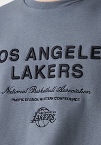 Šedá mikina se černým vyšívaným textem, který zní "Los Angeles Lakers" a "Národní basketbalová asociace." Obsahuje žebrovaný límec.