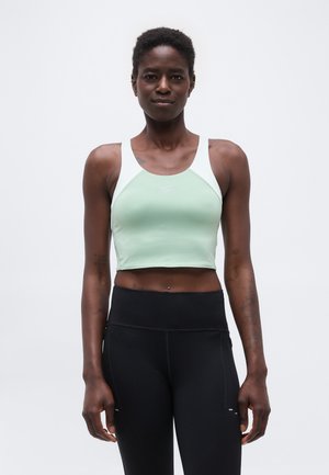 Atletyczna osoba stojąca, ubrana w jasnooliwkowy i biały sportowy top z odsłoniętym brzuchem oraz czarne legginsy z wysokim stanem na jednolitym tle.