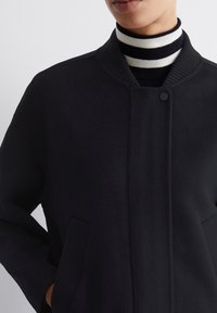 Sort, struktureret frakke med ribbet krave, skjult knaplukning og sidelommer, lagdelt over en sort og hvid stribet turtleneck.