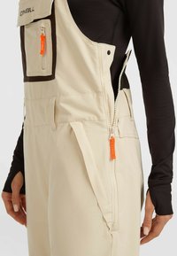 Beige overall met een ritszak met oranje accenten en een zijrits voor gemakkelijke toegang. De stof lijkt glad en duurzaam.