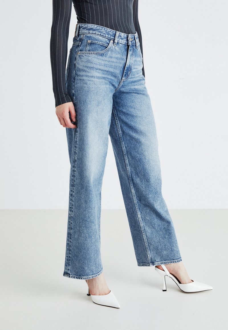 Femme portant un jean droit bleu clair, des talons hauts blancs pointus et un haut noir rayé à manches longues, debout sur un sol clair.