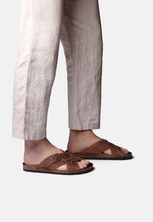 Nohy v hnedých kožených sandáloch so sponou a prekríženými remienkami, v kombinácii so svetlo béžovými ľanovými krátkymi nohavicami na bielom pozadí.