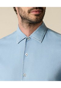 Světle modrá denimová košile s klasickým límcem, dvěma knoflíky a hladkým povrchem. Podrobné prošívání podél švů.