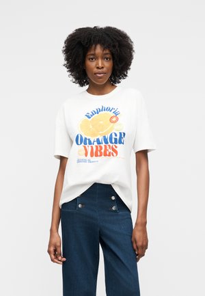 Kvinde med krøllet hår iført en hvid t-shirt med grafik af en appelsinskive og teksten "Euphoria Orange Vibes", kombineret med højtaljede blå bukser.
