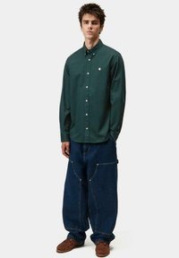 Camicia verde a maniche lunghe con colletto, abbinata a jeans in denim larghi con grandi tasche, completata da scarpe marroni senza lacci.