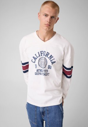 Jeune homme aux cheveux blonds courts portant une chemise blanche à manches longues avec le texte "California Athletics Department" et un jean bleu, sur un fond uni.