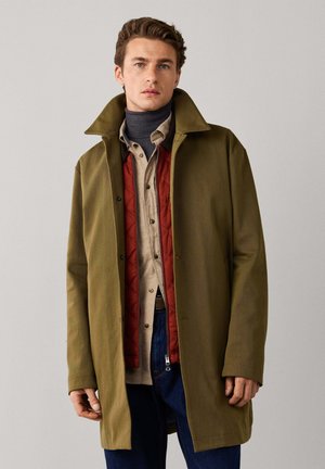 Man wearing layered clothing including a green coat, red quilted vest, beige shirt, and dark turtleneck against a plain background.