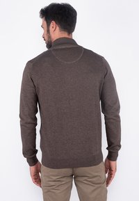 Brun, langermet genser med rund hals, ribbestrikket kant og mansjetter, laget av et mykt materiale, brukt over beige bukser.