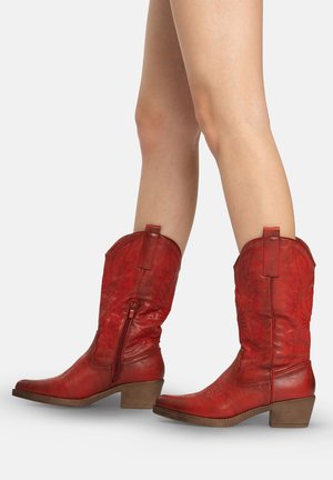 Gambe con stivali da cowboy in pelle rossa fino a metà polpaccio, con dettagli di cuciture e tacchi bassi impilati in legno, mostrati su uno sfondo semplice.