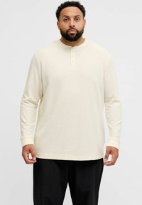 Langermet, kremfarget henley-skjorte med ribbet tekstur, knappedetaljer ved kragen og tettsittende mansjetter. Stylet med mørke bukser.