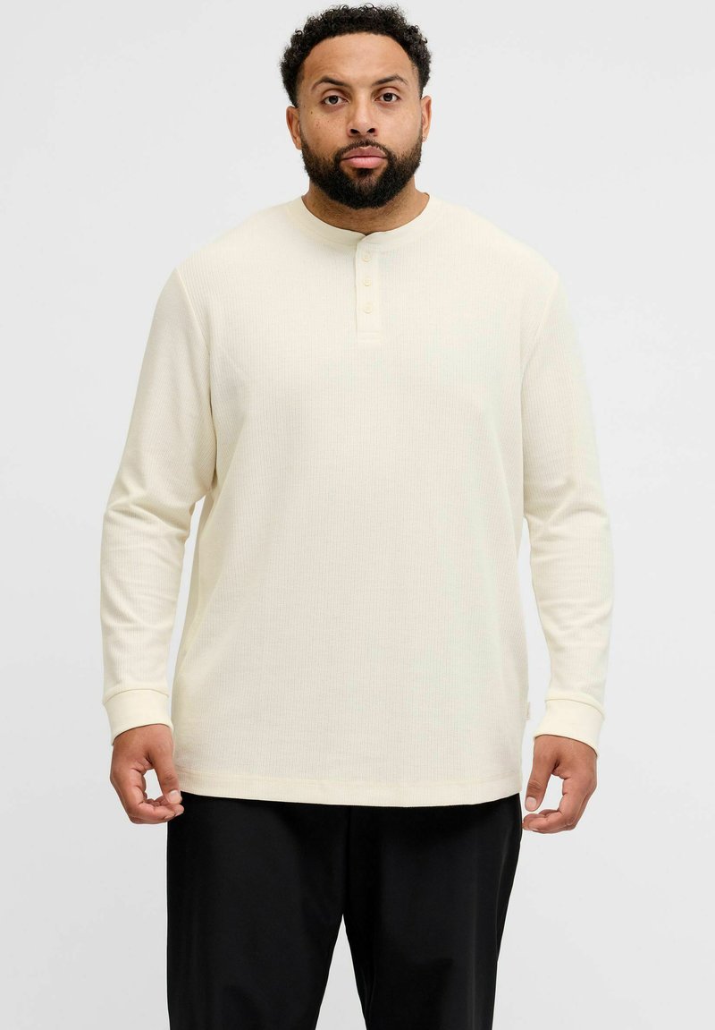 Langermet, kremfarget henley-skjorte med ribbet tekstur, knappedetaljer ved kragen og tettsittende mansjetter. Stylet med mørke bukser.