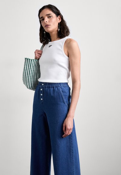 Biała szarfowa bluzka z logo, niebieskie dżinsowe spodnie z szerokimi nogawkami na wysokim stanie oraz torba w zielono-białe paski z guzikami.