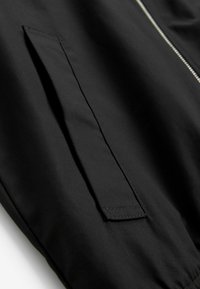 Gros plan d'une poche de veste noire avec rabat et une fermeture éclair argentée visible sur un tissu lisse.