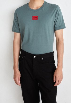 Green cotton t-shirt with short sleeves and a red rectangular patch branded "HUGO" at the chest, worn with black jeans.