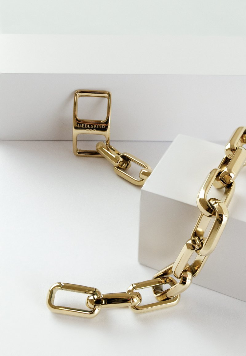 Gouden metalen ketting met ovale schakels, voorzien van een rechthoekig hardware stuk met de gravure "LIEBESKIND," rustend op een witte ondergrond.