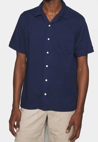Marinblå kortärmad skjorta med knappar, tillverkad av ett lätt material. Den har en bröstficka och klassisk krage, med vita knappar.