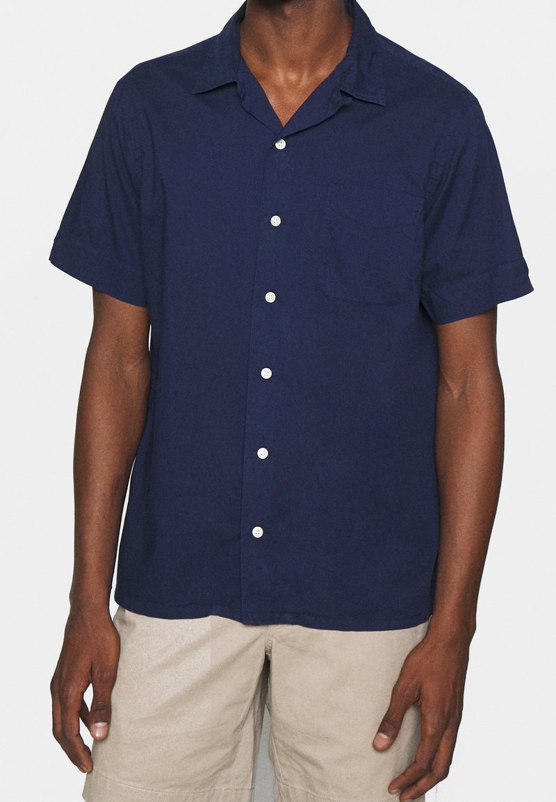 Marinblå kortärmad skjorta med knappar, tillverkad av ett lätt material. Den har en bröstficka och klassisk krage, med vita knappar.