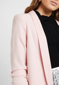 Světle růžový blazer s módním střihem a elegantními liniemi. Disponuje měkkou texturou a otevřeným předním dílem, doplněným černým žebrovaným topem pod ním.