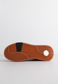 Schwarze Sneaker-Sohle mit ausgeprägtem Zickzackmuster, strukturiertem Gummi für besseren Halt und einem runden weißen Akzentetikett an der Unterseite.