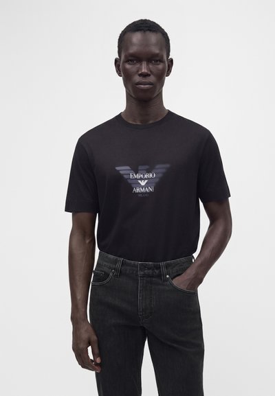 Άνδρας με μαύρο t-shirt Emporio Armani και σκούρα τζιν με το ένα χέρι στην τσέπη πάνω σε απλό λευκό φόντο.