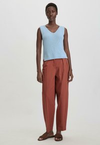 Valk bleu sans manches en tricot avec un col en V, associé à un pantalon structuré couleur rouille et des sandales à glisser marron.