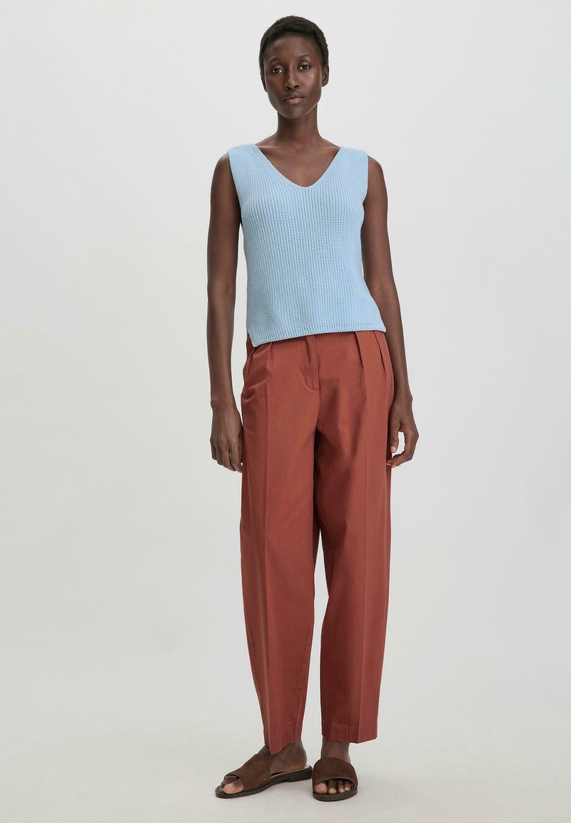 Valk bleu sans manches en tricot avec un col en V, associé à un pantalon structuré couleur rouille et des sandales à glisser marron.