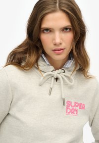 Grijze hoodie met verstelbare aantrekkoord, voorzien van zilverkleurige hardware en een roze "SUPERDRY" logo. Gelaagd over een lichtblauw overhemd met kraag.