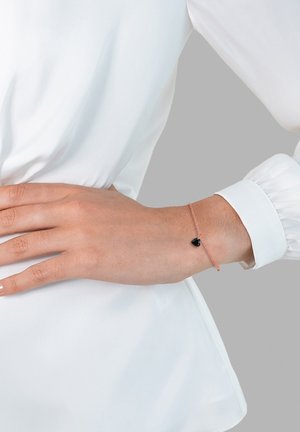 Rose gold chain bracelet with a heart-shaped black pendant, worn on a hand resting against a white blouse. Smooth texture and delicate design.