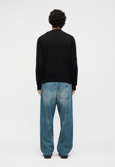 Homme aux cheveux bouclés portant une chemise noire à manches longues et un jean bleu ample, debout de dos sur un fond blanc uni.