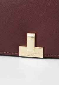 Primo piano di una borsa in pelle rosso scuro con una fibbia rettangolare in oro incisa con "ALDO" su una superficie bianca.