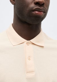Gros plan sur une personne portant un polo beige clair avec une patte de boutonnage à deux boutons et un col plat.