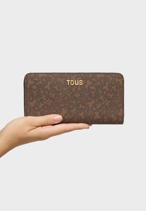 Main tenant un portefeuille TOUS marron à motifs avec des lettres dorées sur un fond gris clair uni.