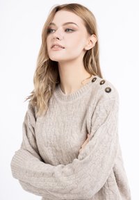 Beige kötött pulóver, kerek nyakkivágással, vastag bordás mandzsettákkal és három díszgombbal a bal vállon. Puha textúra.