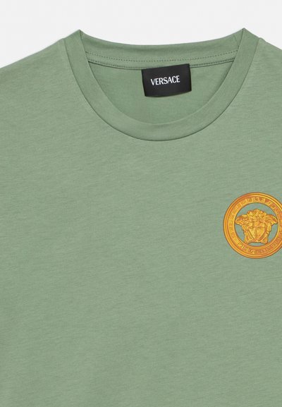 Ανοιχτό πράσινο T-shirt με στρογγυλή λαιμόκοψη, με μικρό πορτοκαλί και χρυσό έμβλημα Medusa στο αριστερό στήθος και ετικέτα Versace στο εσωτερικό του γιακά.