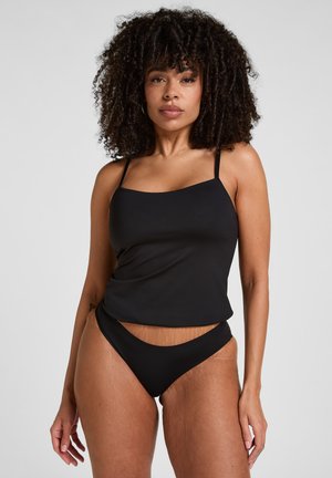 Frau mit lockigem Haar, die ein schwarzes ärmelloses Top und passende hochgeschnittene Bikinihöschen vor einem schlichten hellen Hintergrund trägt.