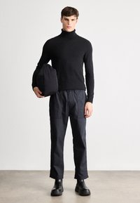 Suéter preto de gola alta combinado com calças pretas de cintura elástica. A modelo segura uma mala preta, usando botas de cano curto pretas. Design minimalista.