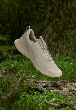 Halványbarna, könnyű terepfutócipő, texturált tapadós talppal, zöld, mohás talaj és apró kövek fölött lebeg egy erdei környezetben.