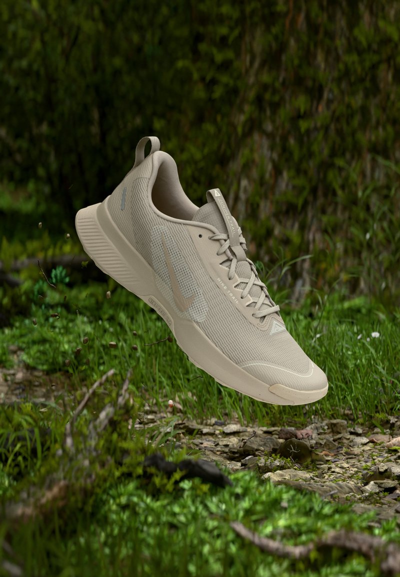 Béžová ľahká bežecká topánka na trase s textúrovanou podrážkou, zavesená nad zeleným machovým povrchom a malými kameňmi v lesnom prostredí.