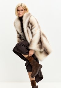 Donna bionda che indossa una gonna a righe, stivali marroni con calze spesse e un ampio cappotto di pelliccia, in posa contro uno sfondo bianco semplice.