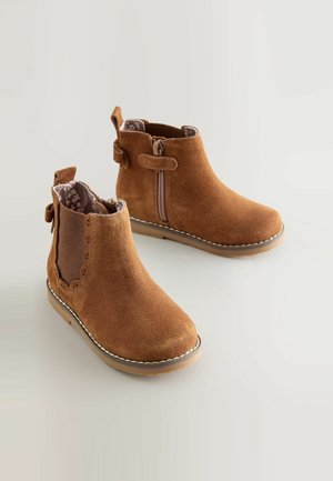 Bottines en suède marron avec une fermeture éclaire et des panneaux latéraux élastiques. Présentent un bord festonné sur le dessus et une doublure texturée à l'intérieur.